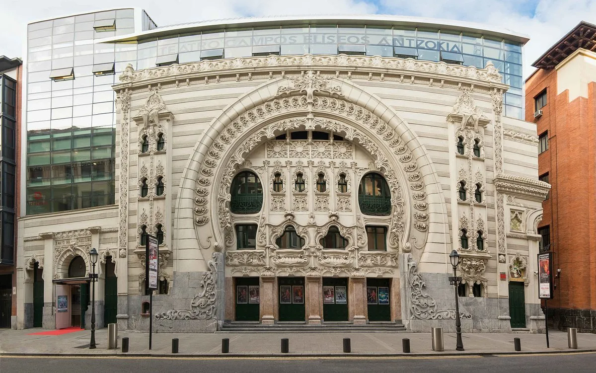 De sierlijke witte art-nouveaugevel van Teatro Campos Elíseos Antzokia met boogramen en decoratief metselwerk, geflankeerd door moderne gebouwen van glas en baksteen.