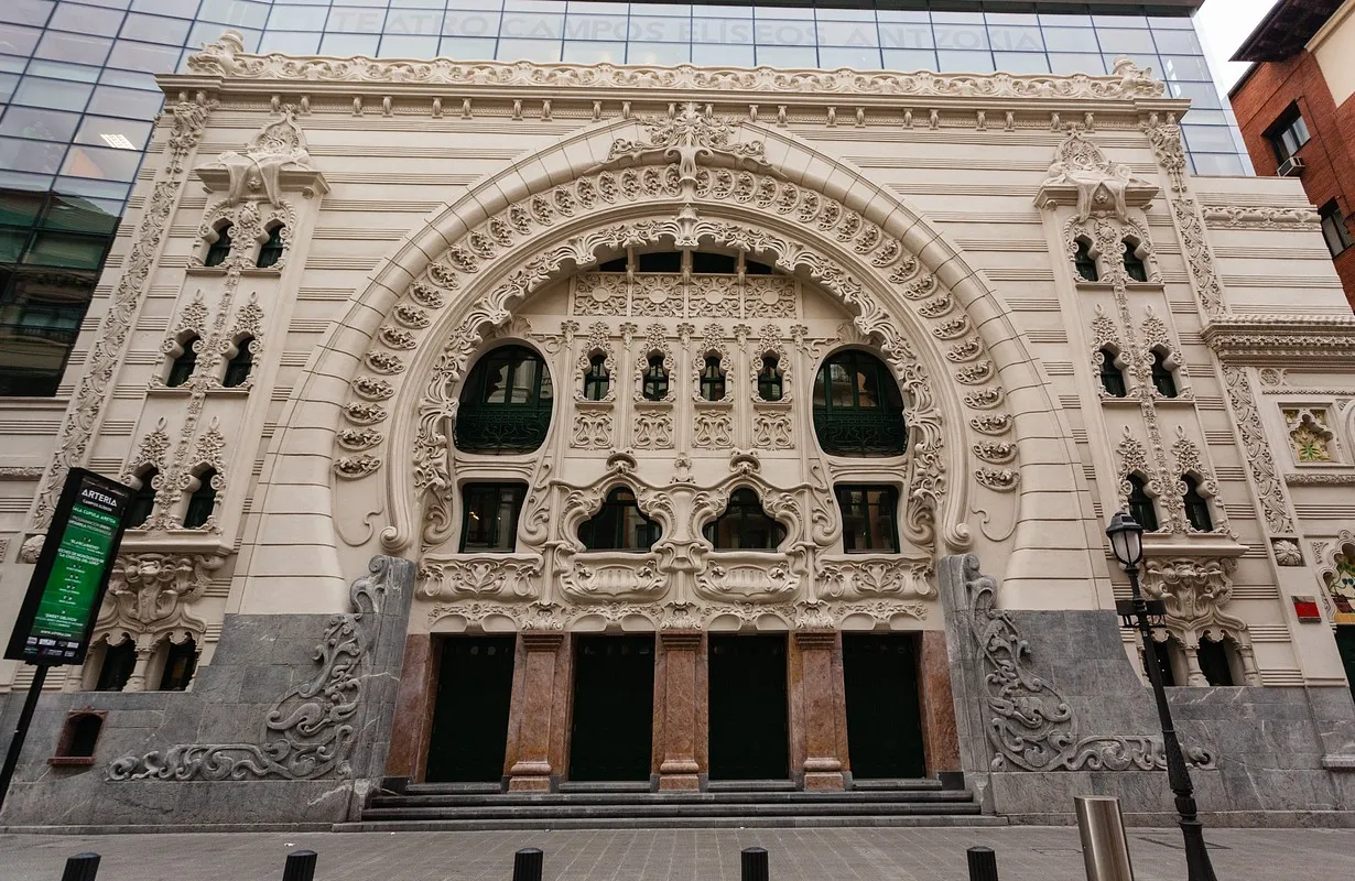 Sierlijke witte art-nouveaugevel van een theater met uitgebreid beeldhouwwerk in steen, boogvensters, decoratieve reliëfs en een modern glazen gebouw zichtbaar op de achtergrond.