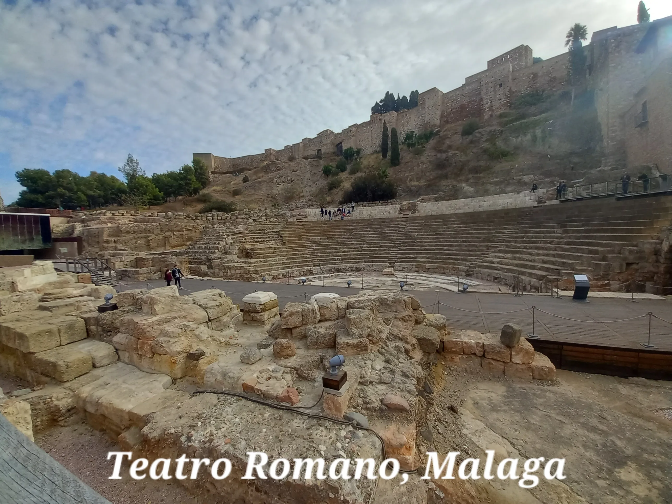Szeroki widok na ruiny Teatru Rzymskiego w Maladze, ukazujący kamienne rzędy siedzeń oraz twierdzę Alcazaba na wzgórzu powyżej, z napisem 'Teatro Romano, Malaga' u dołu.