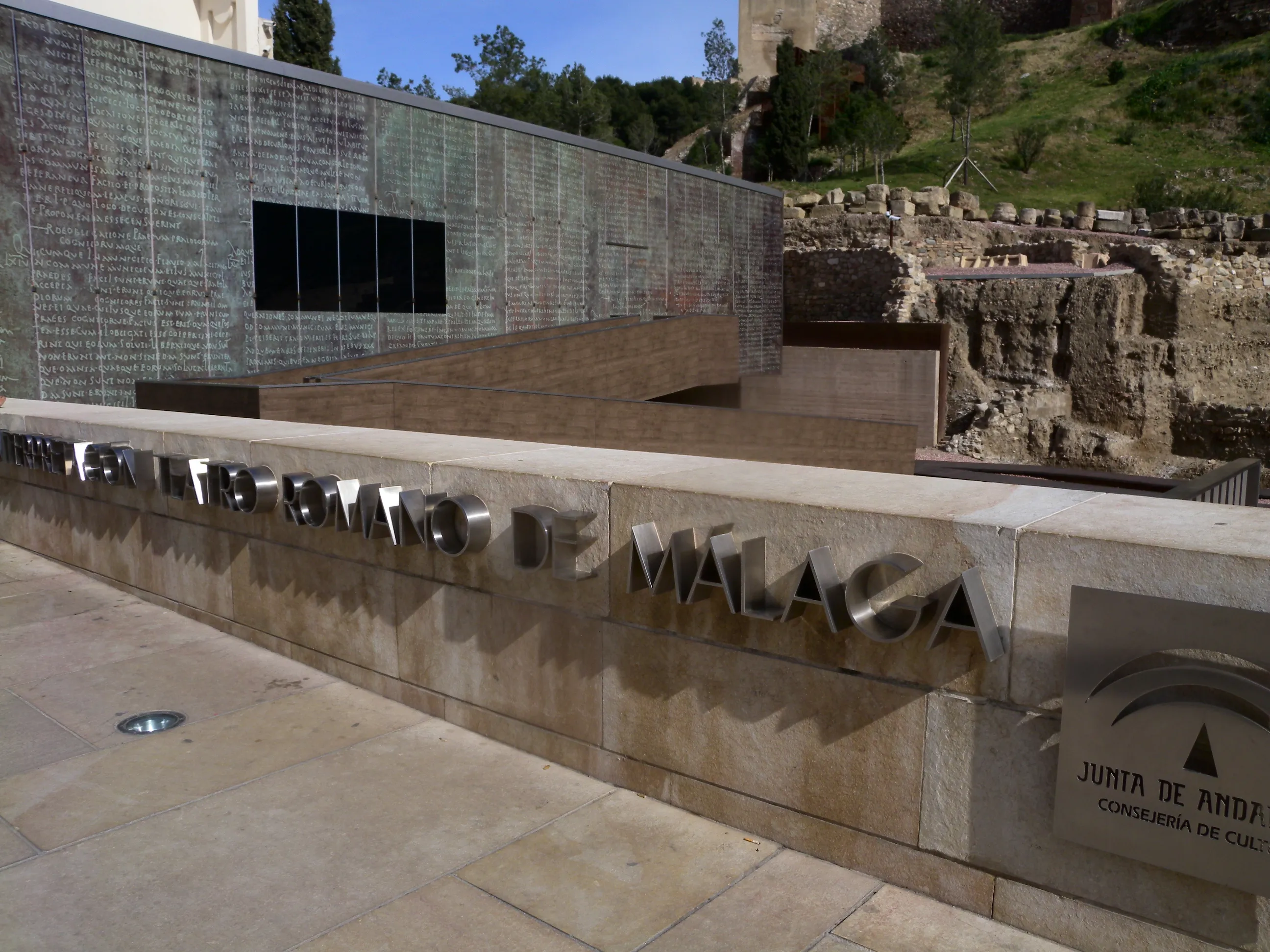 Edificio moderno con fachada de cristal y muro de piedra con la inscripción 'TEATRO ROMANO DE MÁLAGA' en la entrada, con las antiguas ruinas romanas y la colina de la Alcazaba al fondo.