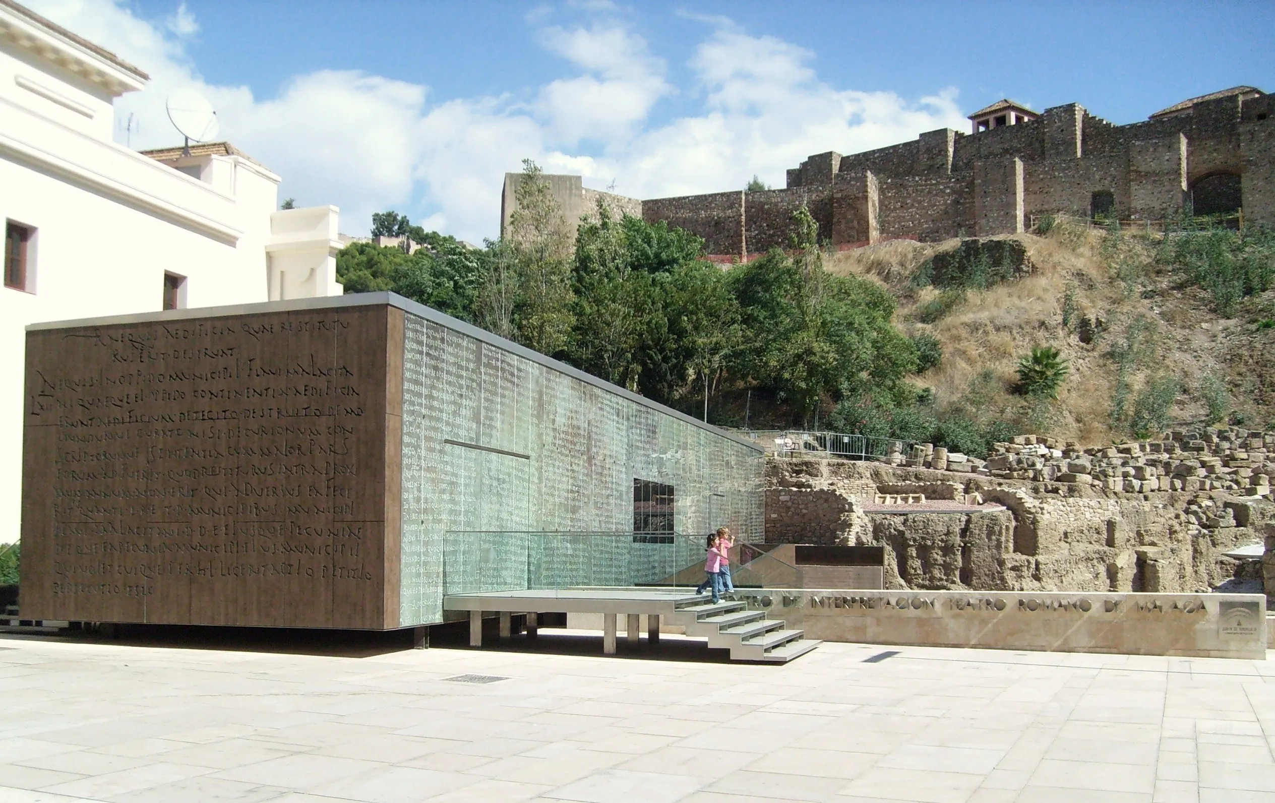 Moderno centro de interpretación de cristal y piedra junto a las antiguas ruinas del teatro romano, con la fortaleza de la Alcazaba sobre la colina al fondo.