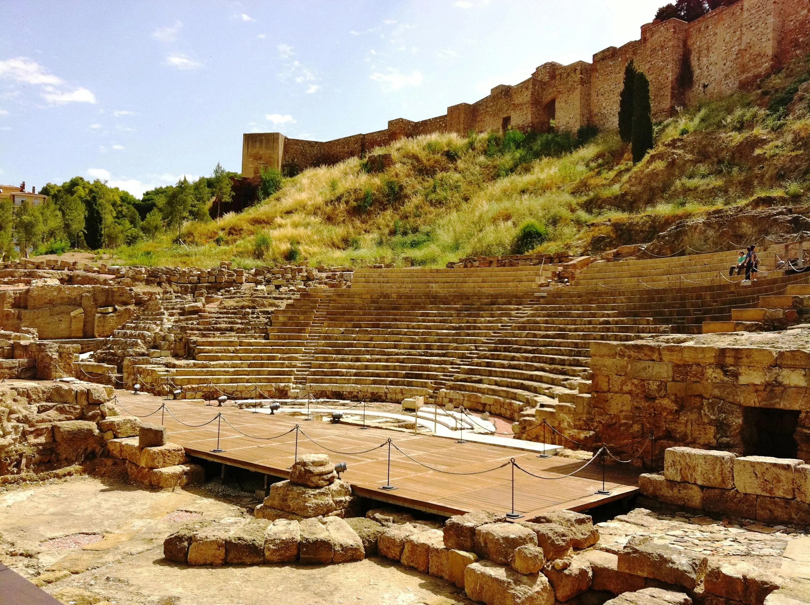 Vista panorámica de las ruinas del teatro romano de Málaga, que muestra las gradas de piedra y una plataforma de escenario de madera con las murallas de la Alcazaba en la colina superior.