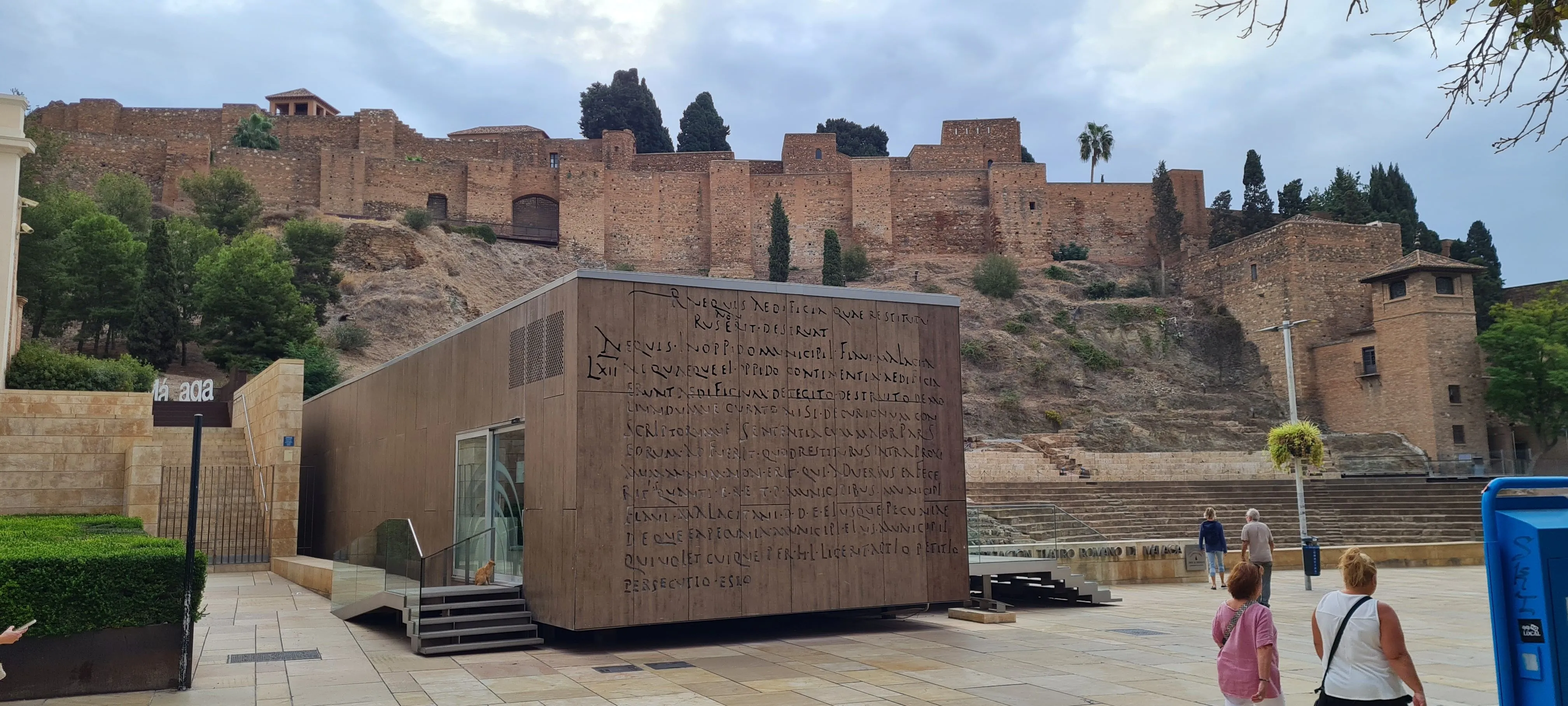 Vista panorámica exterior del Teatro Romano de Málaga que muestra el moderno edificio rectangular del centro de interpretación con fachada inscrita, las ruinas del antiguo teatro romano y las murallas de la alcazaba en la colina al fondo