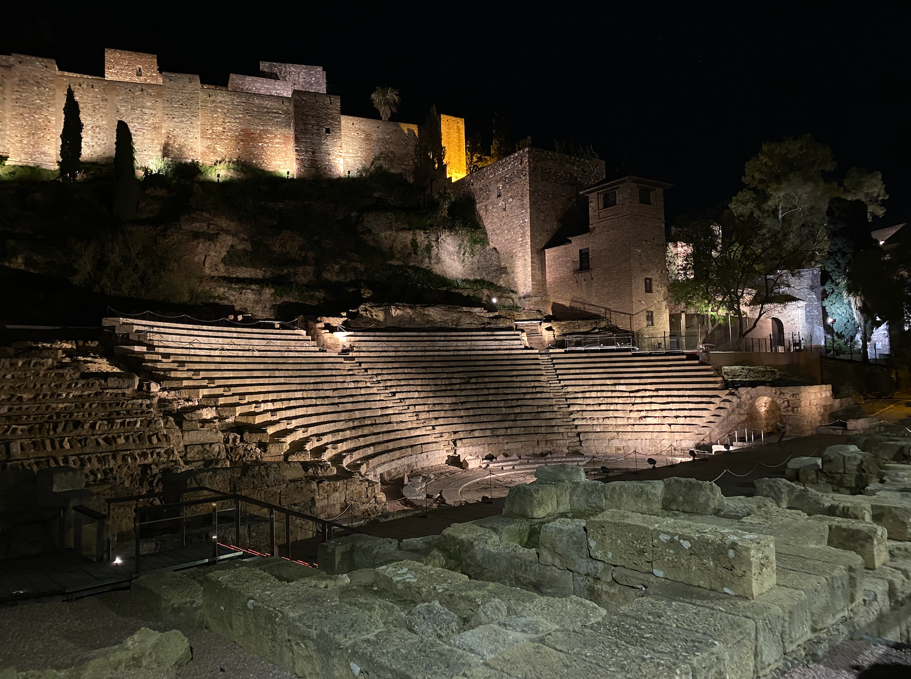 Vista nocturna de las gradas de piedra iluminadas del teatro romano, con las murallas de la fortaleza de la Alcazaba iluminadas en la colina superior.