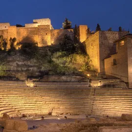 Asientos de piedra iluminados del Teatro Romano de Málaga al atardecer, con la alcazaba iluminada en la colina que se alza sobre él.