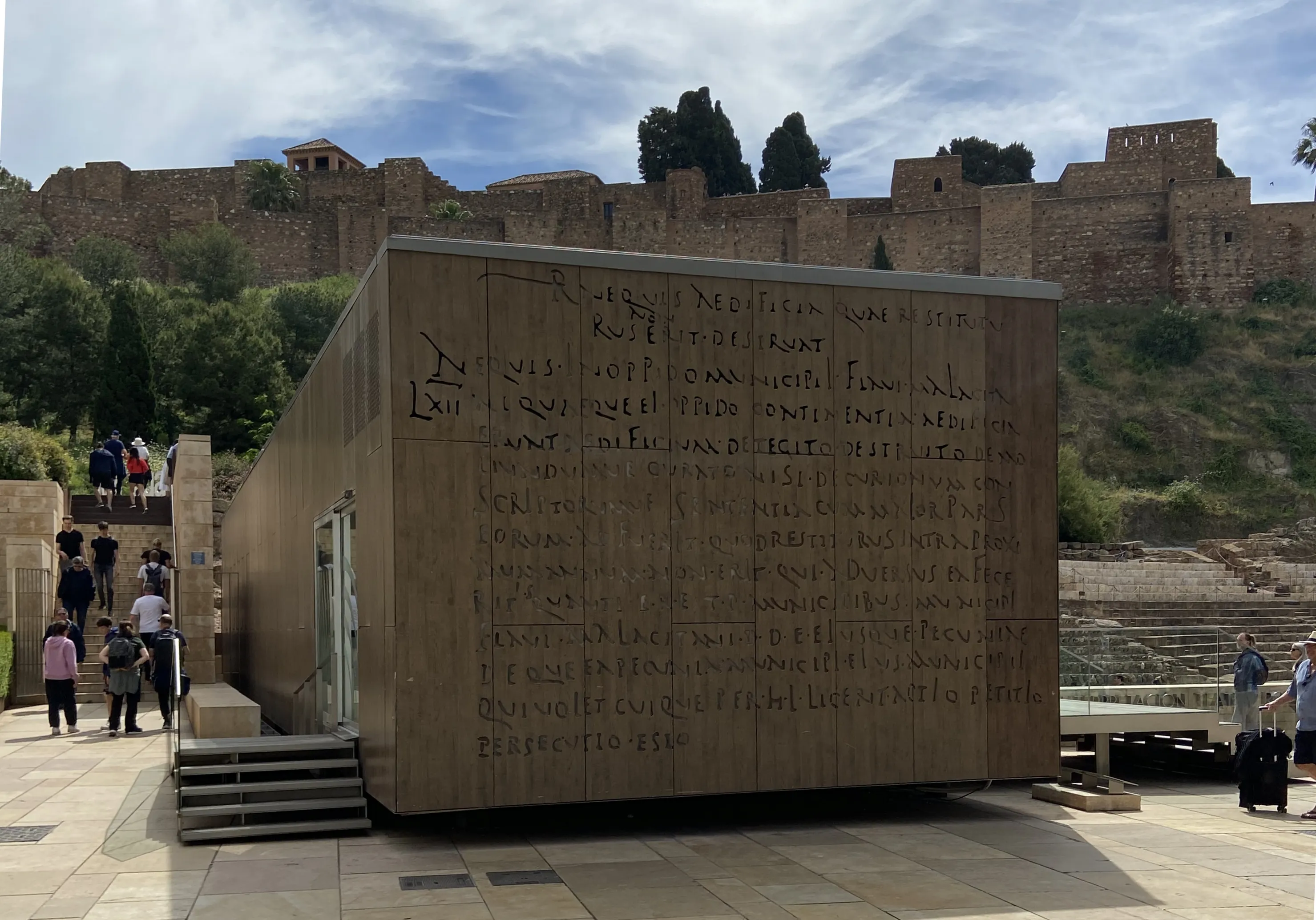 Edificio de centro de interpretación revestido de piedra de planta rectangular con inscripciones latinas talladas en su fachada, con las murallas de la fortaleza de la Alcazaba visibles en la ladera detrás y visitantes caminando cerca.