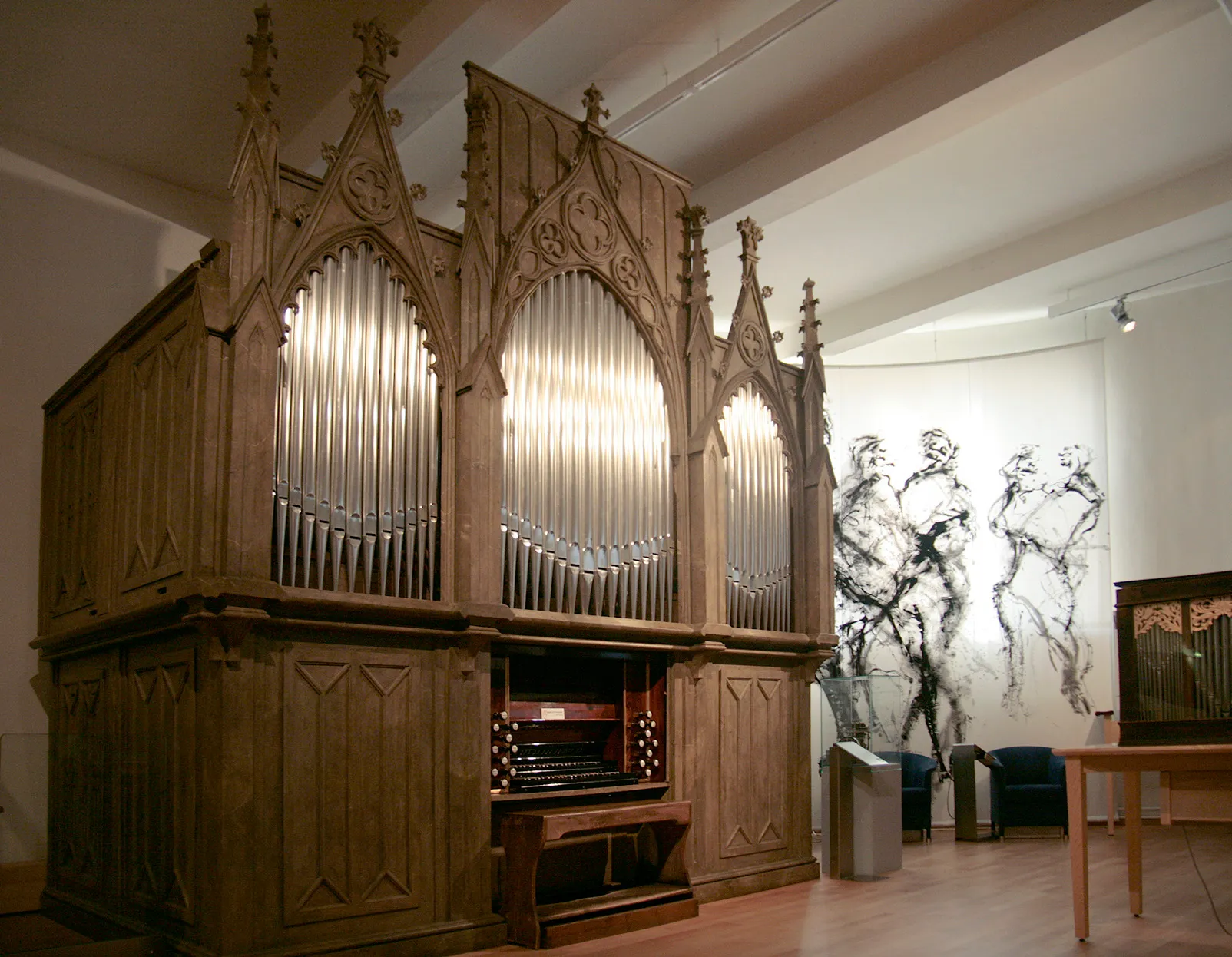 Een grote houten pijporgel met gotische snijwerken en verlichte pijpen, binnen tentoongesteld naast een muurschildering.