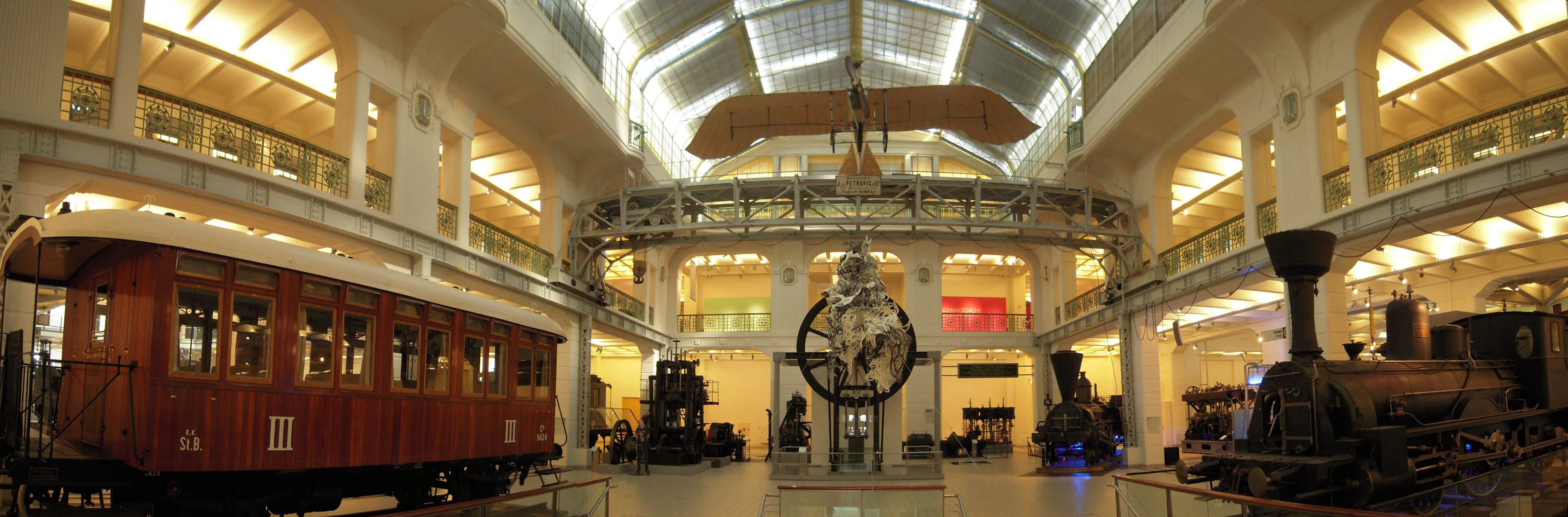 Interior de un salón de museo con un vagón de tren rojo, una locomotora de vapor y un avión suspendido del techo