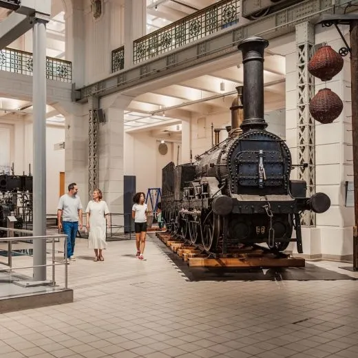 Una locomotora de vapor negra expuesta en un salón de museo con tres personas caminando al fondo.