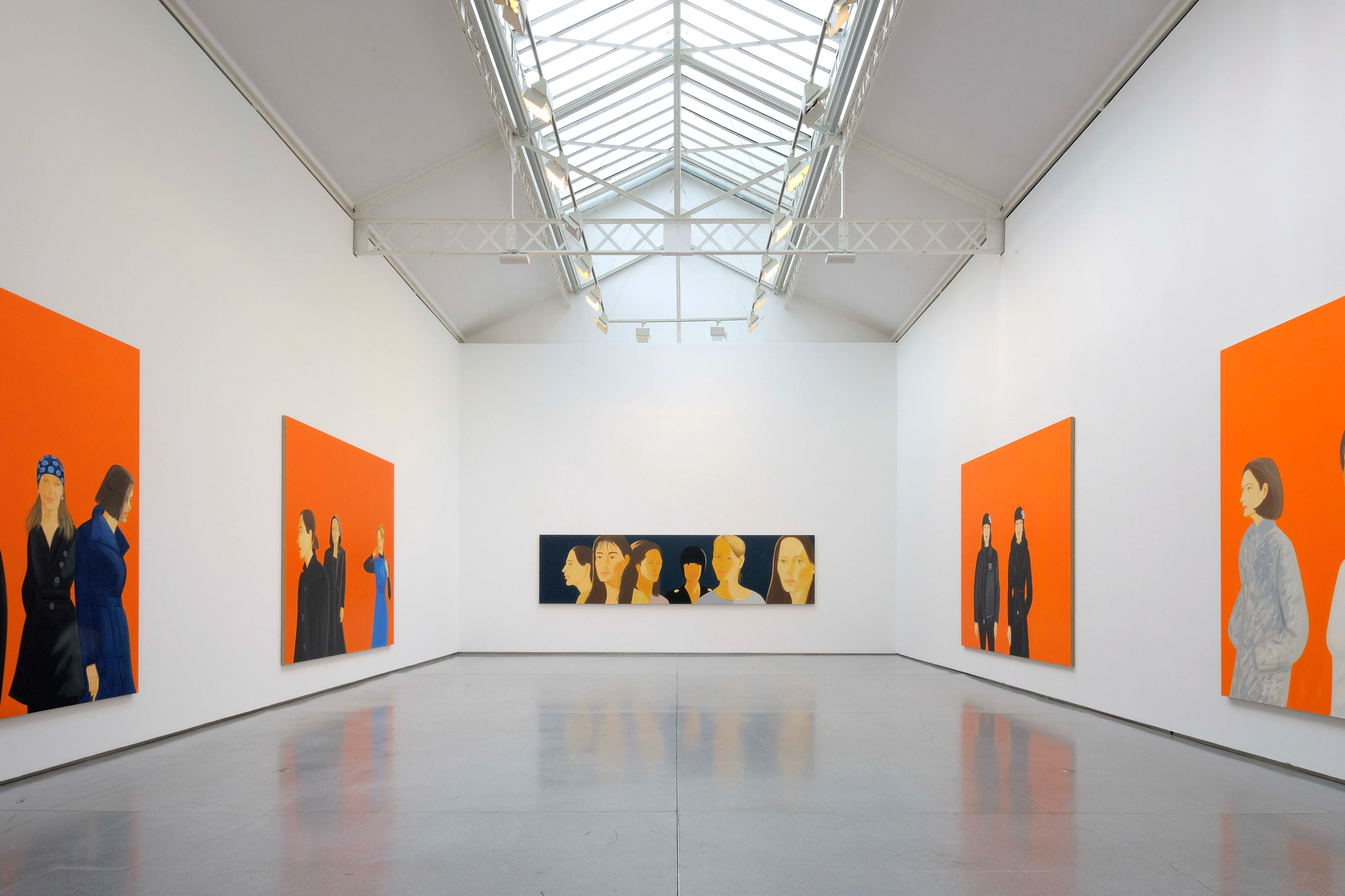 Interior de una galería con grandes pinturas de figuras sobre fondos de color naranja brillante expuestas en paredes blancas bajo un techo de cristal.