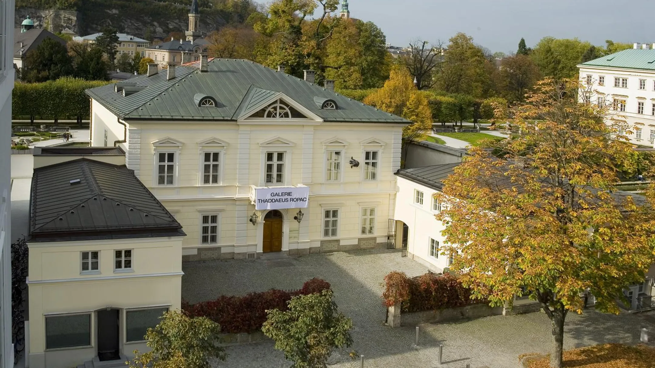 Vista aérea de una villa neoclásica blanca con patio, árboles de otoño y una pancarta de Thaddaeus Ropac en la fachada