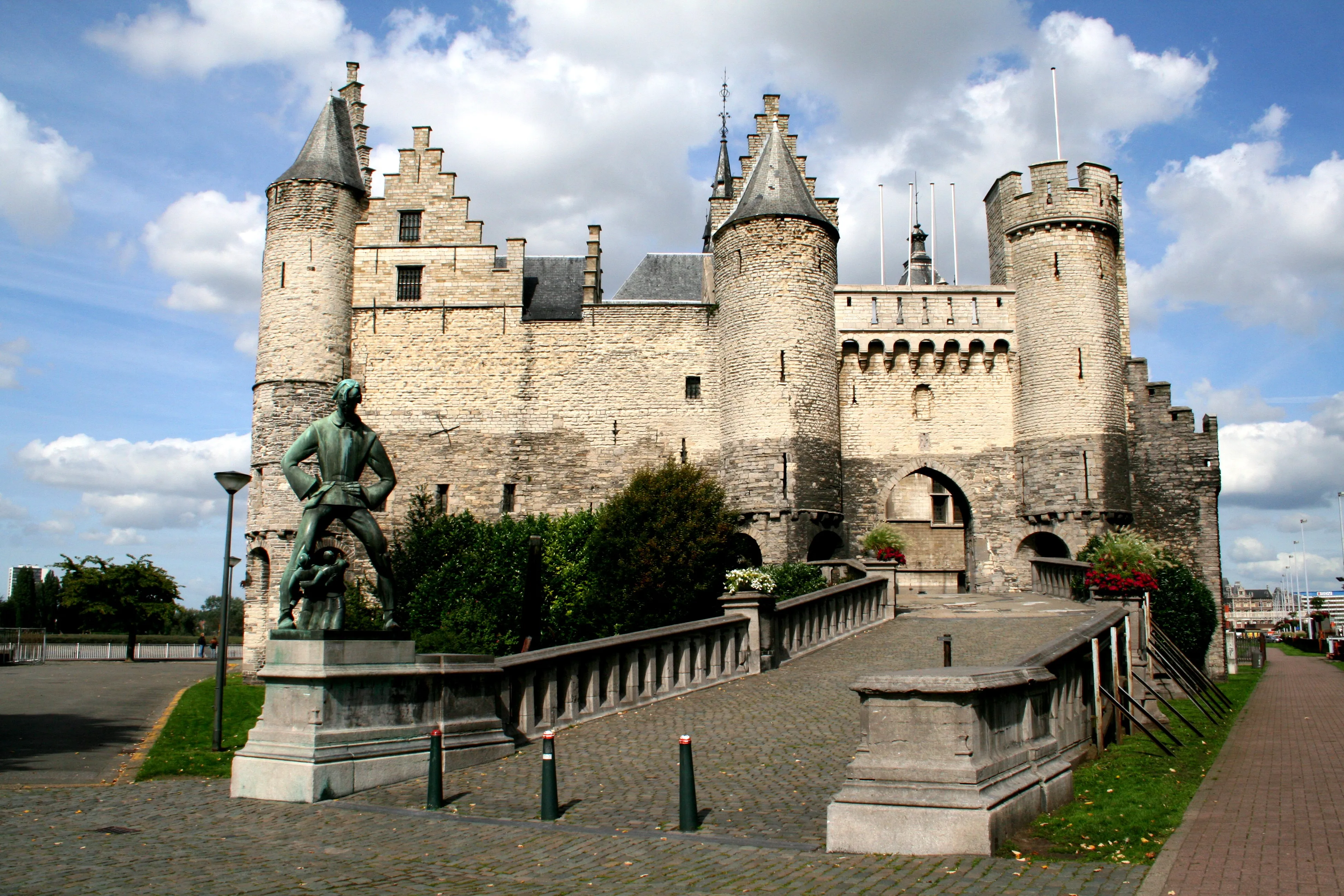 Castillo medieval de piedra con torres redondas y hastiales escalonados, con una estatua de bronce sobre un pedestal en primer plano y un puente de piedra que conduce a una puerta arqueada bajo un cielo azul con nubes