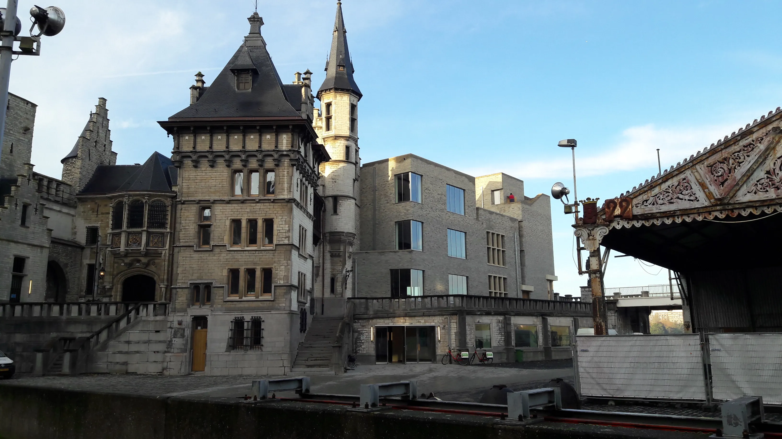 Vista exterior del castillo Het Steen en Amberes con su distintiva torre de piedra y aguja, el edificio moderno adyacente y el paseo marítimo bajo un cielo azul