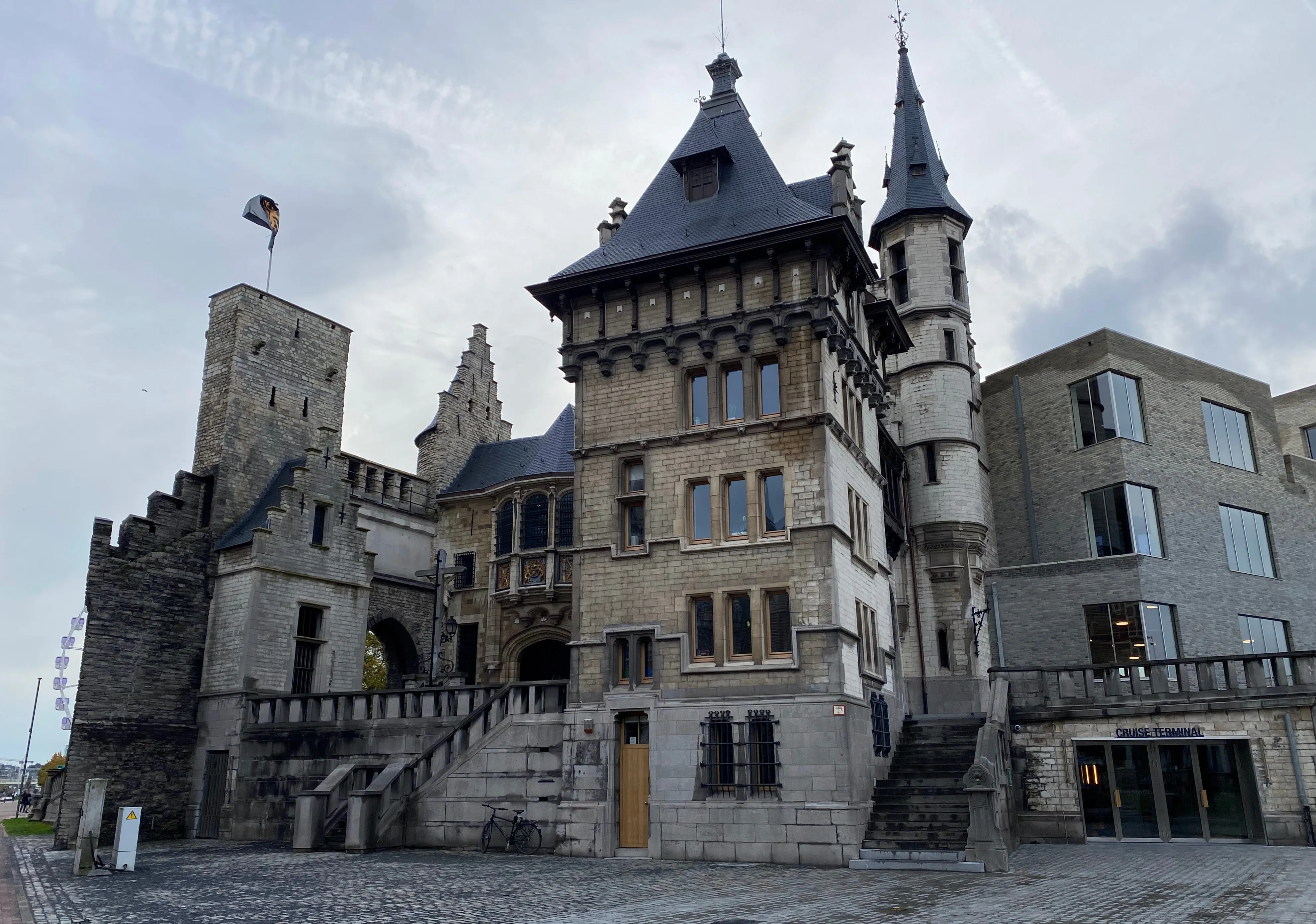 Exterior de un castillo medieval de piedra con múltiples torres, techos de pizarra puntiagudos, entrada arqueada y patio empedrado bajo un cielo nublado