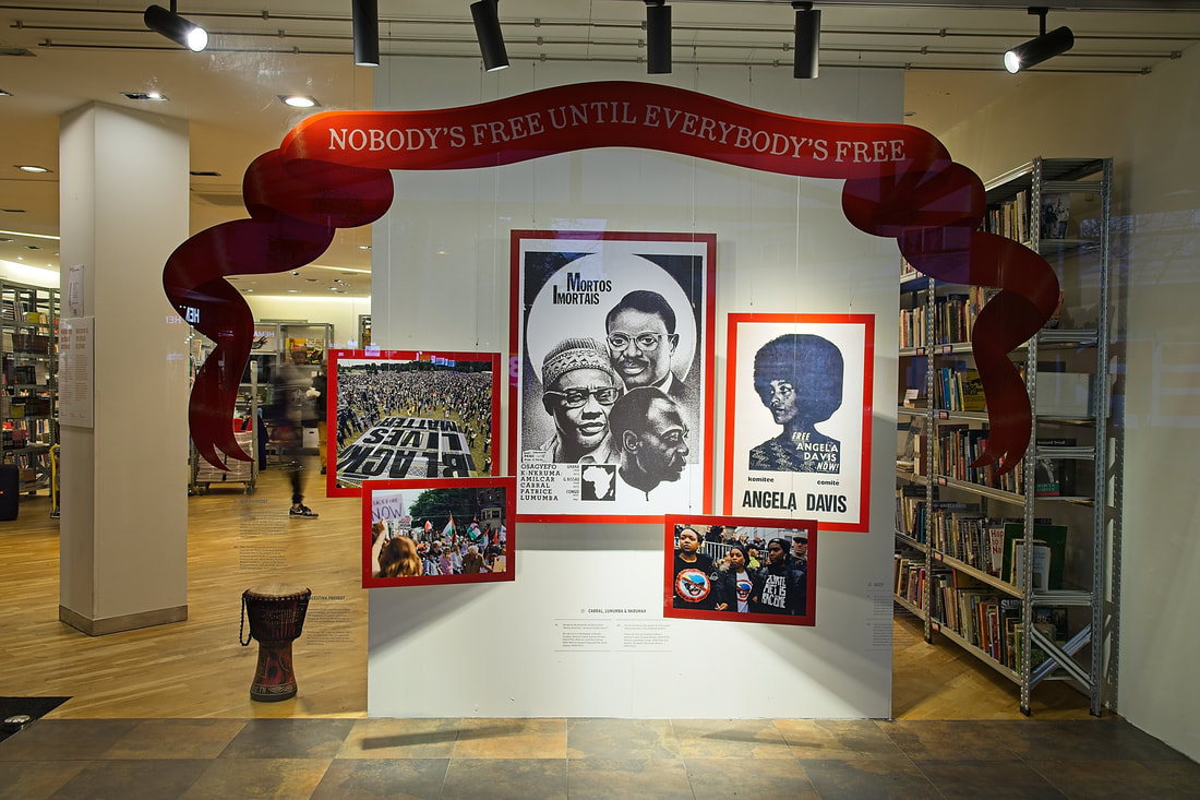 Ściana wystawowa z czerwonym banerem z napisem 'NIKT NIE JEST WOLNY, DOPÓKI WSZYSCY NIE BĘDĄ WOLNI', oprawione plakaty, w tym Angeli Davis, oraz bęben w przestrzeni bibliotecznej z półkami na książki.