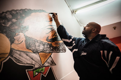 Mężczyzna maluje barwny mural przedstawiający osobę w chuście na głowie i z logo Nike na wewnętrznej ścianie