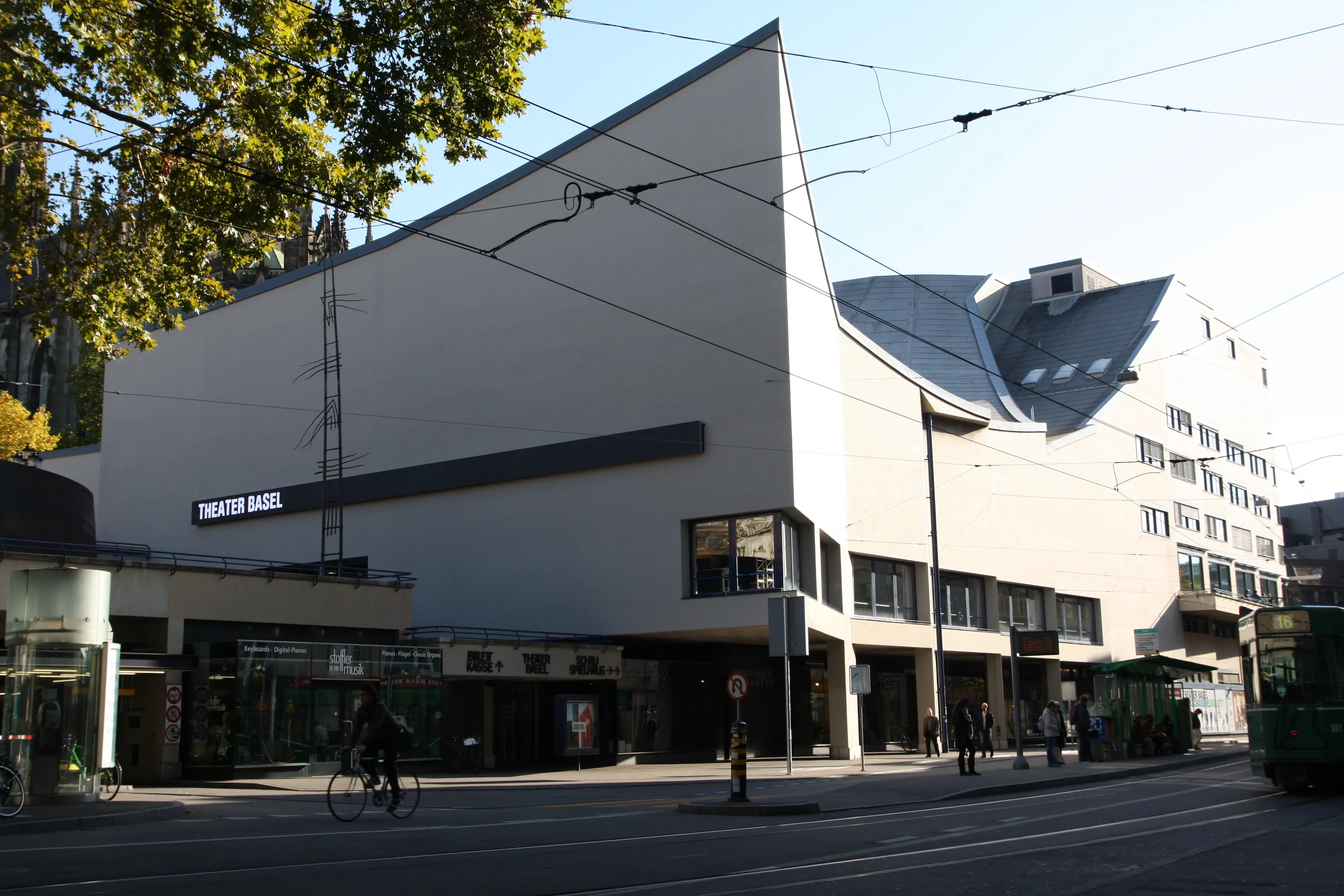 Widok z zewnątrz na Theater Basel, ukazujący białą, kanciastą fasadę budynku z szyldem teatru, linie tramwajowe nad ulicą oraz ulicę z pieszymi i autobusem.
