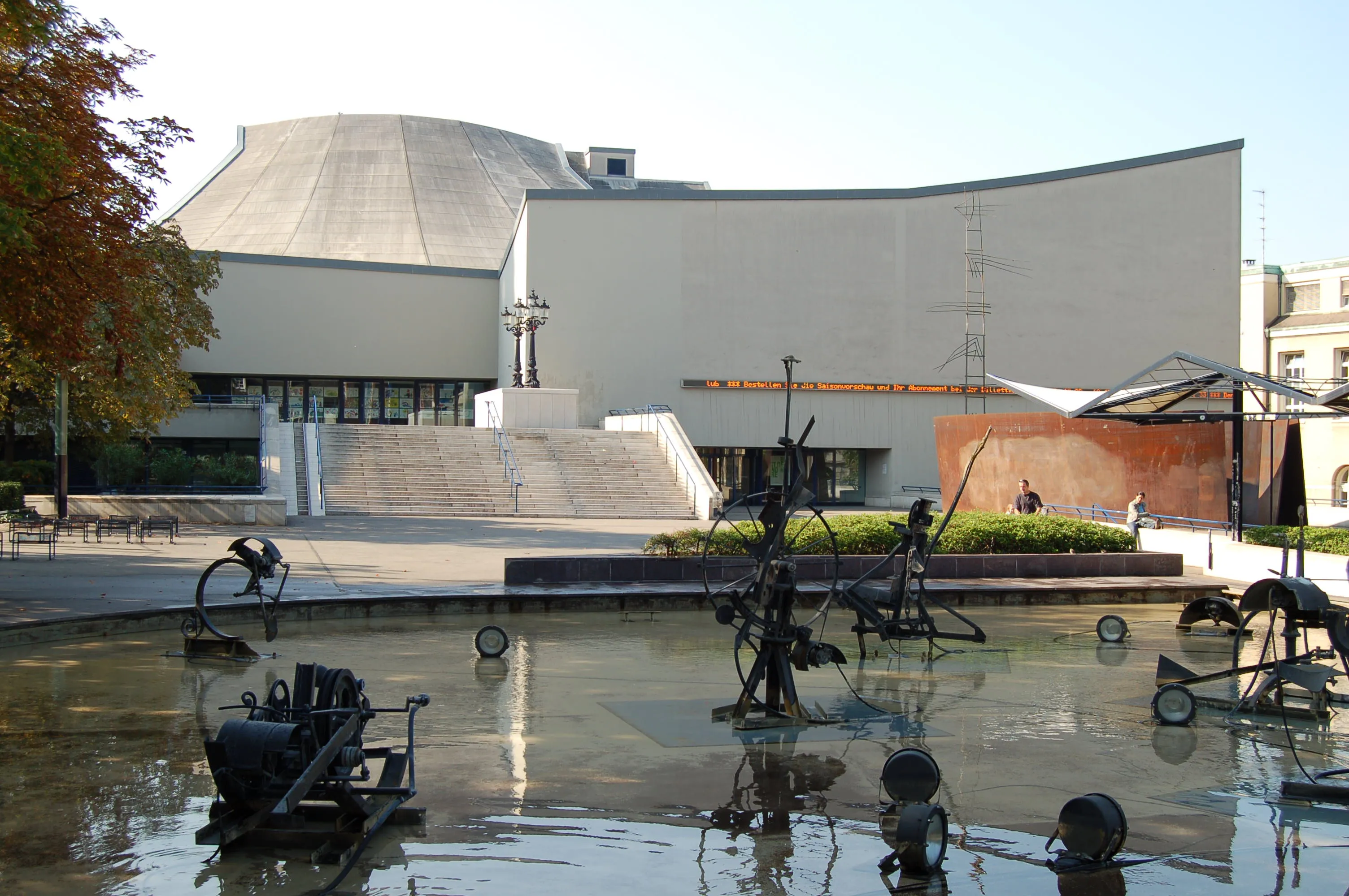 Biała, zakrzywiona fasada Theater Basel z kopułą w tle, widziana znad fontanny z mechanicznymi metalowymi rzeźbami
