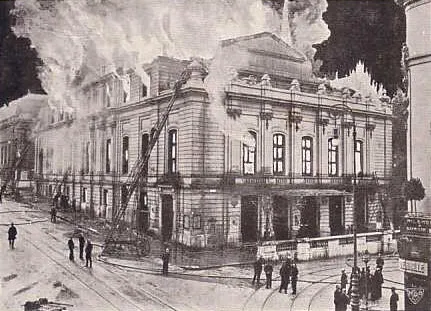 Historyczne czarno-białe zdjęcie budynku Theater Basel ogarniętego płomieniami i gęstym dymem podczas pożaru w 1904 roku, z widocznymi na ulicy drabinami strażackimi i ludźmi
