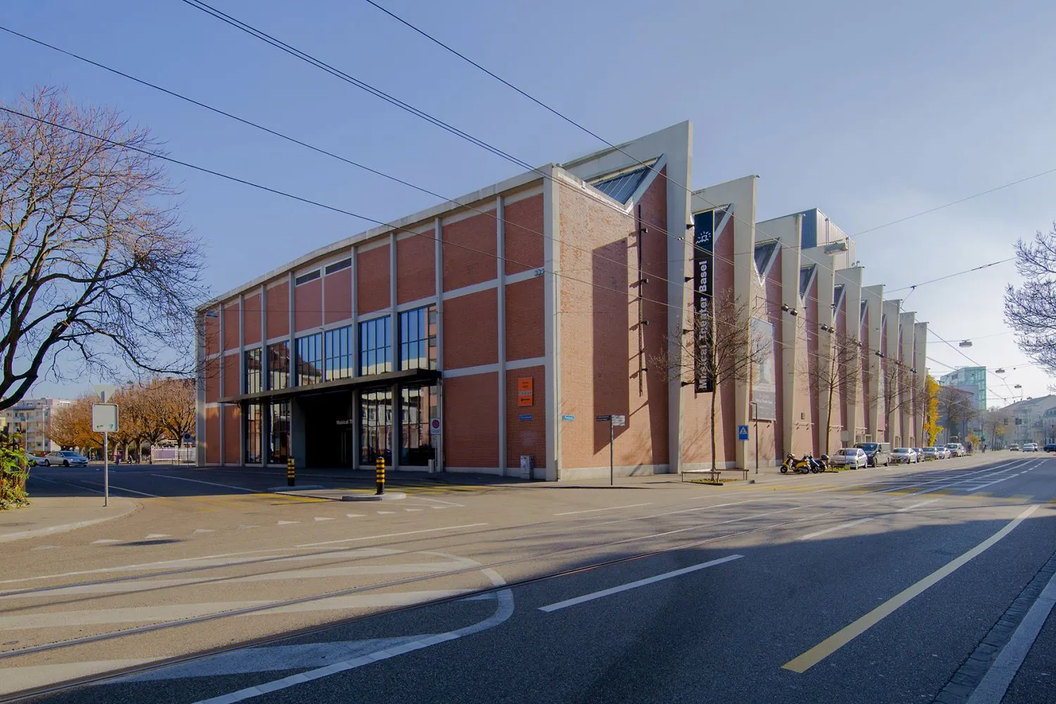 Widok z zewnątrz na Theater Basel, duży budynek z czerwonej cegły z nowoczesną fasadą i szklanym wejściem, widziany z drugiej strony ulicy z torami tramwajowymi.