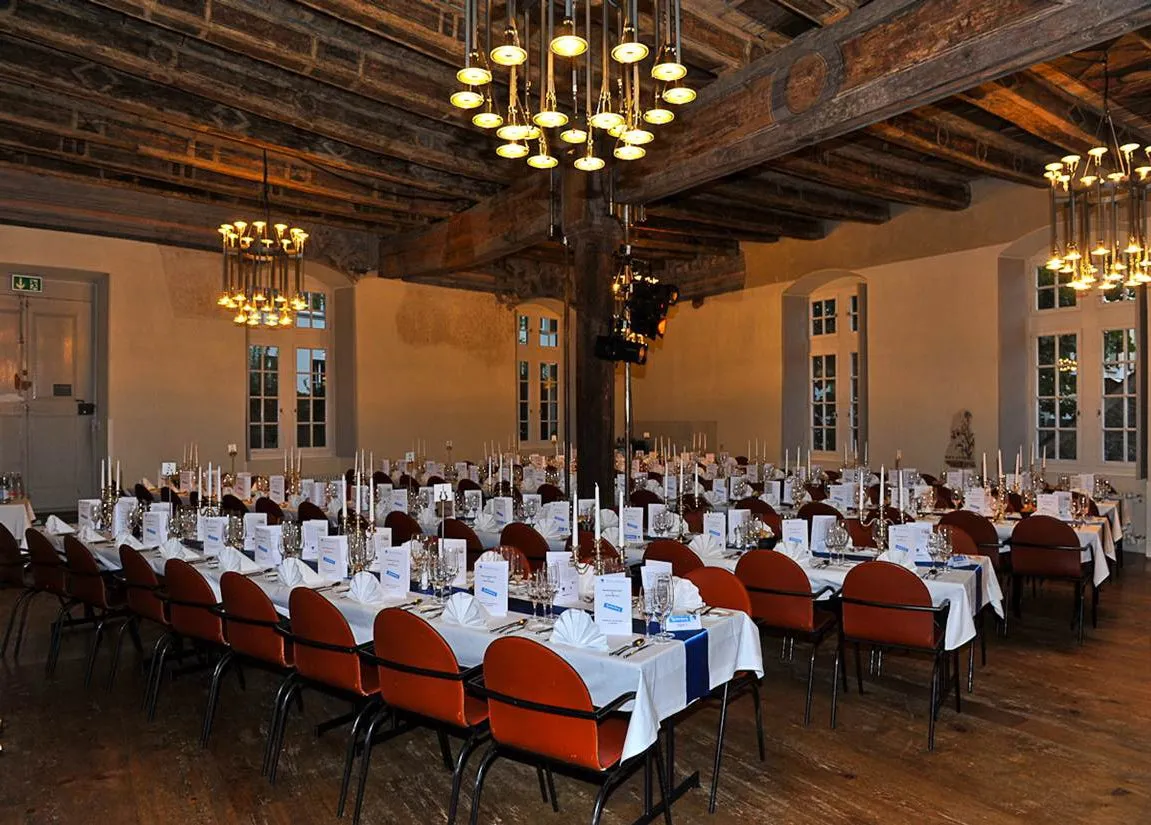 Interieur van een zaal met lange tafels bedekt met witte tafelkleden, rood gestoffeerde stoelen en sierlijke kroonluchters die aan een plafond met zichtbare houten balken hangen