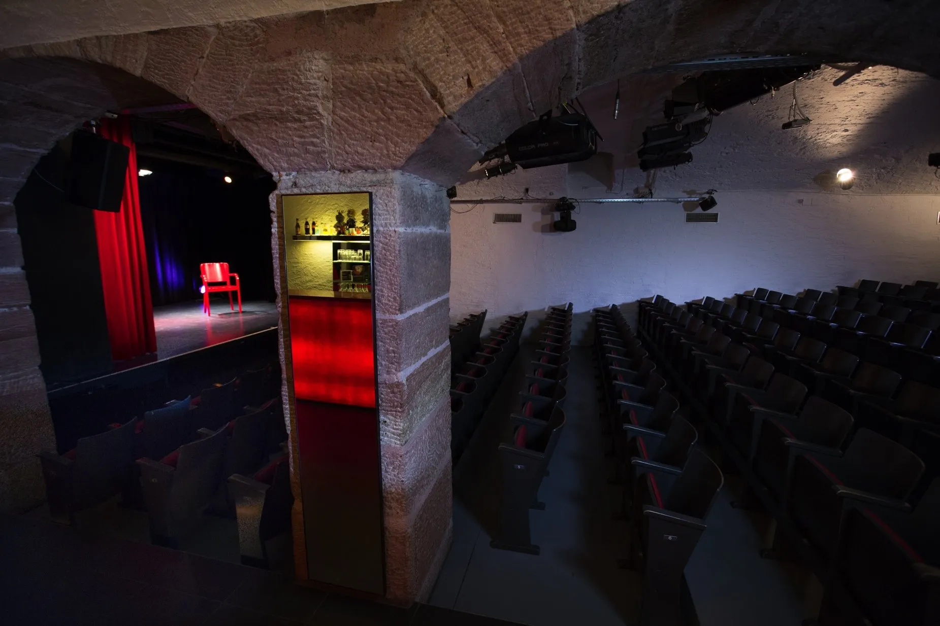 Interieur van Theater Fauteuil met stenen gewelfde bogen, rijen donkere stoelen en een toneelgedeelte met rode gordijnen en een rode stoel