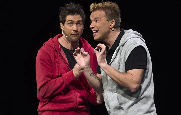 Twee mannelijke artiesten op het podium met headsetmicrofoons, een in een rode hoodie en een in een grijs vest, die geanimeerd gebaren maken tegen een zwarte achtergrond.