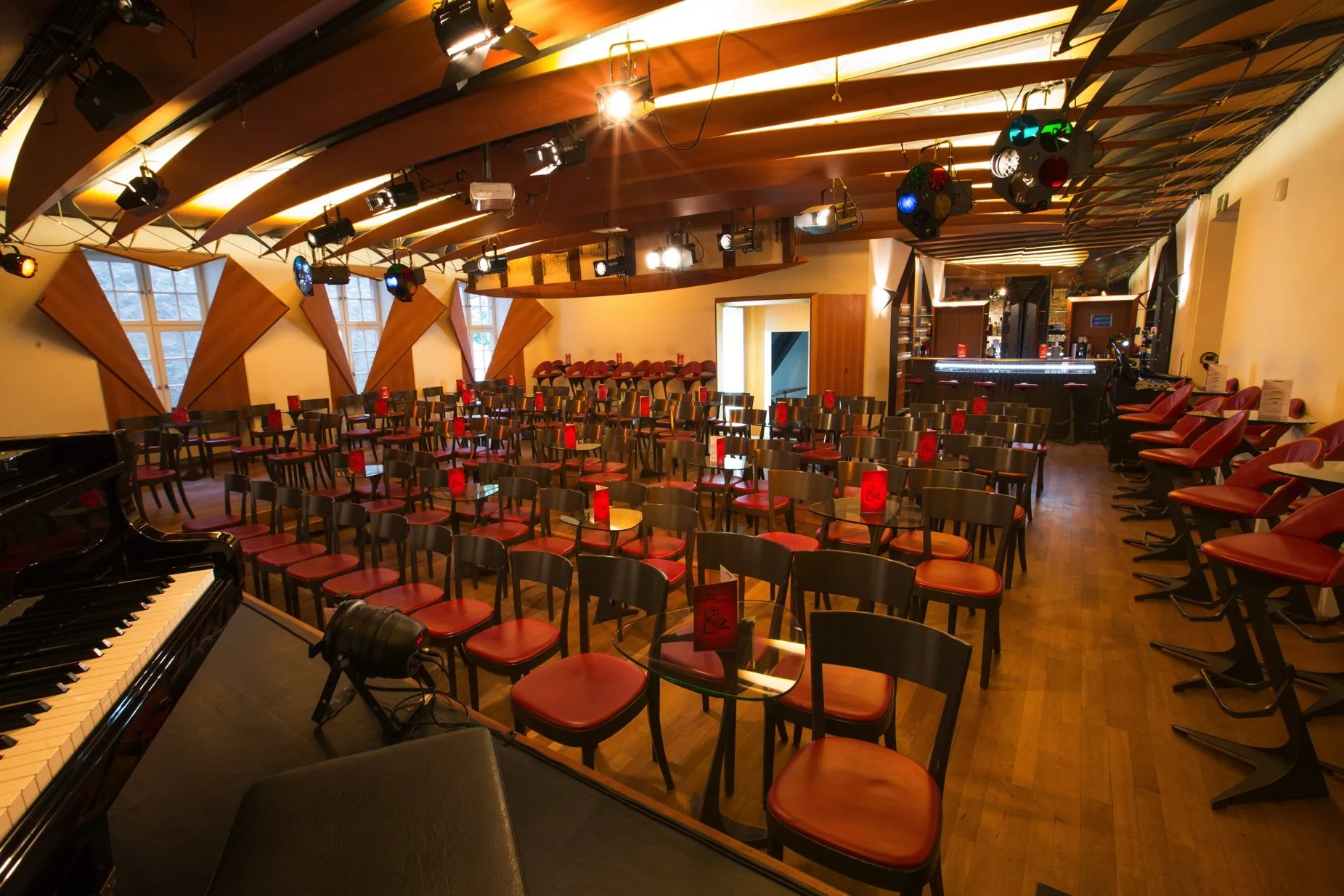 Interieur van een cabaret-theater met rijen houten stoelen met rode kussens, een vleugel op de voorgrond en podiumverlichting gemonteerd aan een houten plafond.