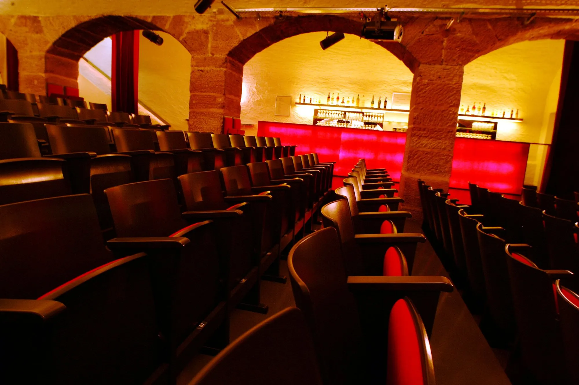 Rijen donkere theaterstoelen in een gewelfde bakstenen ruimte met een rood verlichte bar op de achtergrond