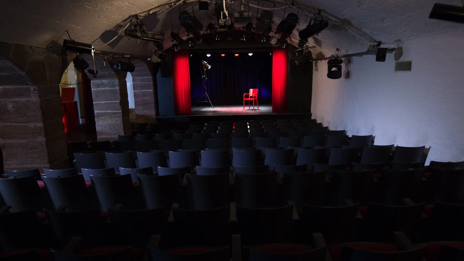Interieur van een theaterzaal met rijen donkere stoelen gericht op een podium dat verlicht is met rood licht, met één enkele rode stoel in het midden