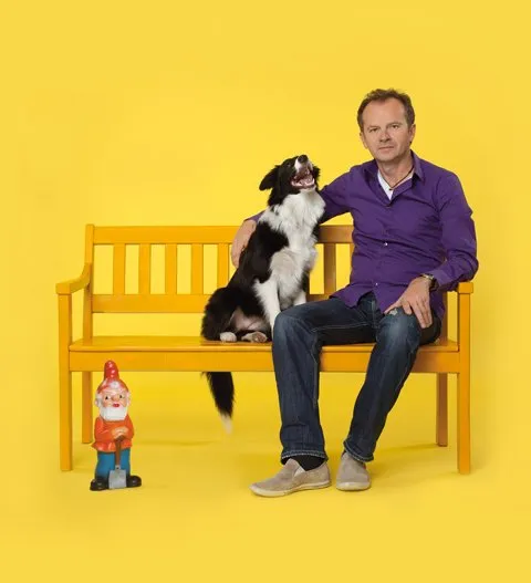 Een man in een paars shirt zittend op een gele bank met een zwart-witte hond, naast een tuinkabouterfiguurtje, tegen een felgele achtergrond.
