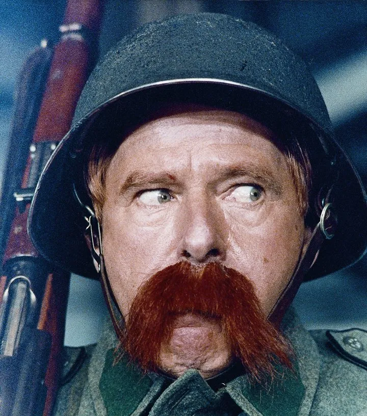 Close-up van een mannelijke acteur met een grote rode snor, die een militaire helm draagt en een geweer vasthoudt.