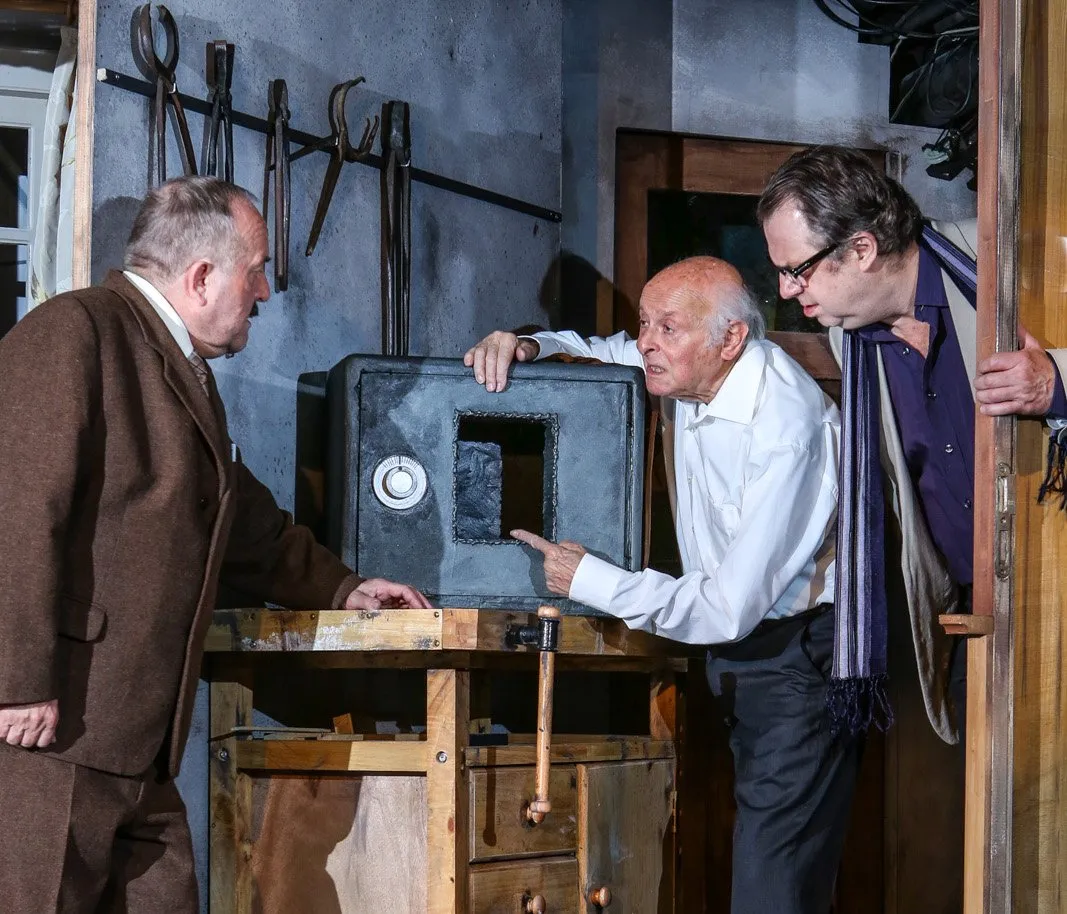 Drie acteurs op een theaterpodium verzameld rond een kluis op een werkbank, met metalen gereedschap aan de muur achter hen.