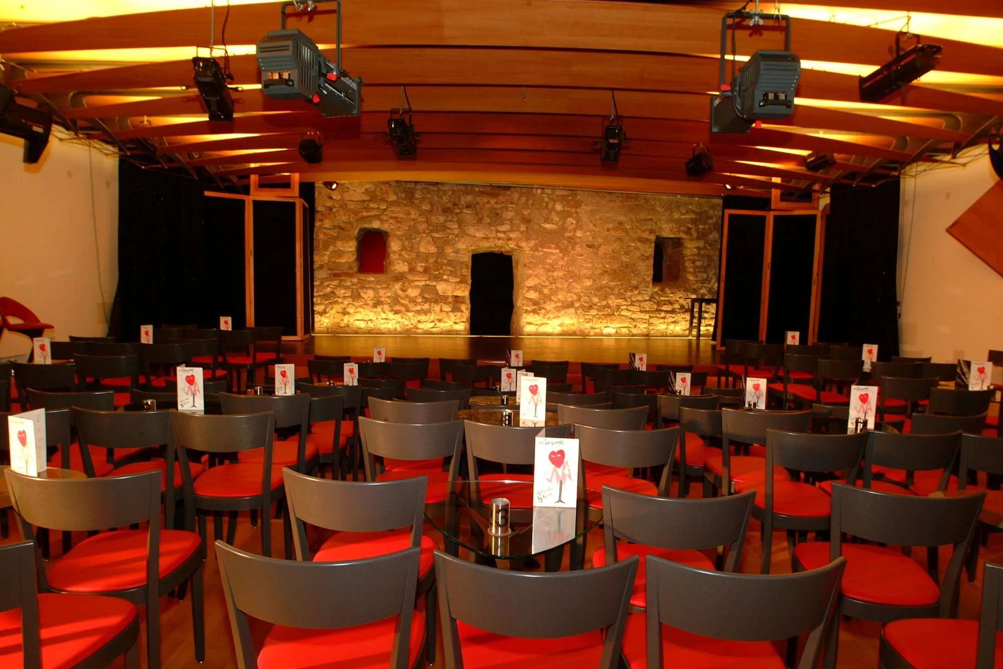 Interieur van een klein cabarettheater met rijen donkere stoelen voorzien van rode kussens, opgesteld rond kleine ronde tafels, gericht op een stenen achtergrond op het podium onder een houten plafond met gemonteerde verlichtingsapparatuur.