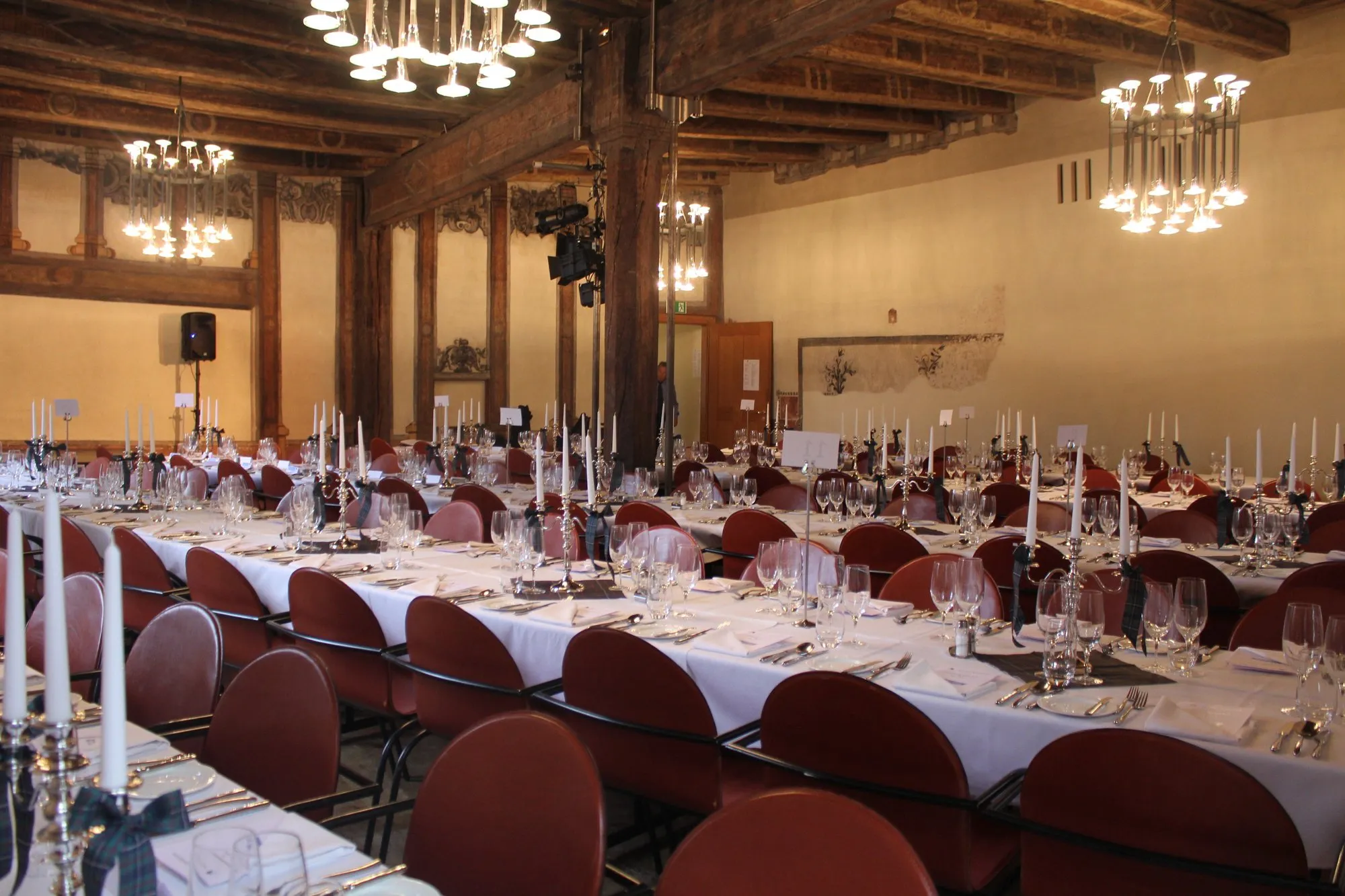 Een grote feestzaal met lange tafels bedekt met witte tafelkleden, rijen rode stoelen, wijnglazen en hoge witte kaarsen, onder plafonds met zichtbare houten balken en meerdere kroonluchters.