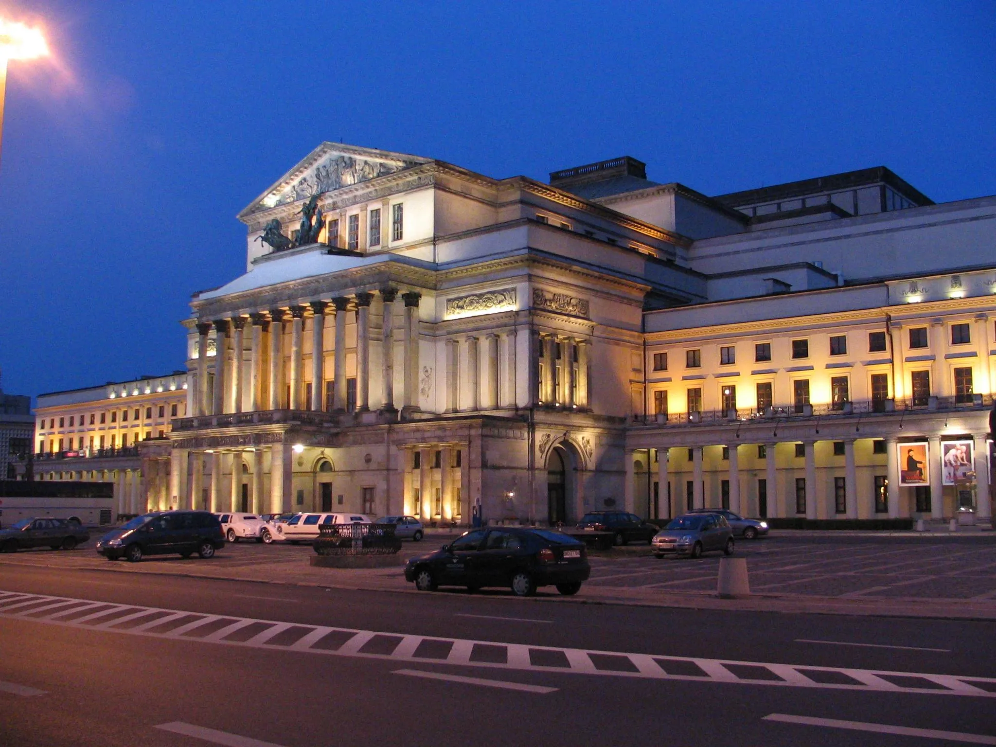 Neoclassical building with columns and a pediment, illuminated at night, with cars on the street in front