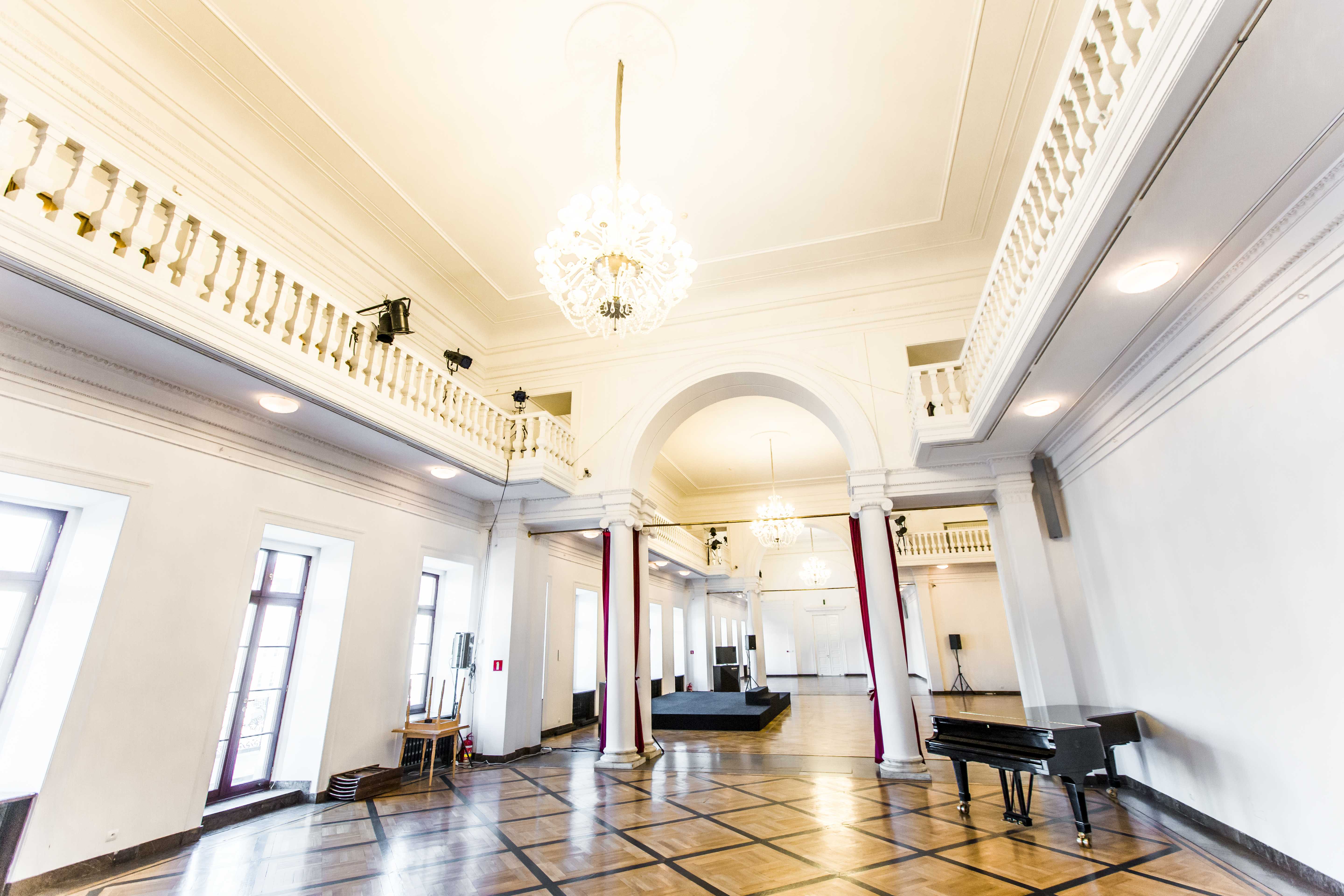Spacious interior hall with high ceilings, white balustrades, arched doorways, and a polished tiled floor. A grand piano is in the foreground and a chandelier hangs from the ceiling.