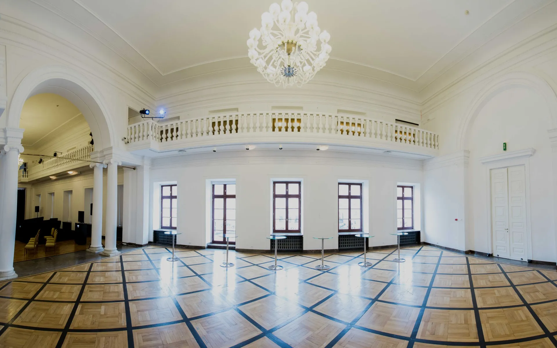 Large hall with white walls, arched doorways, a chandelier, and a wooden floor with a geometric pattern