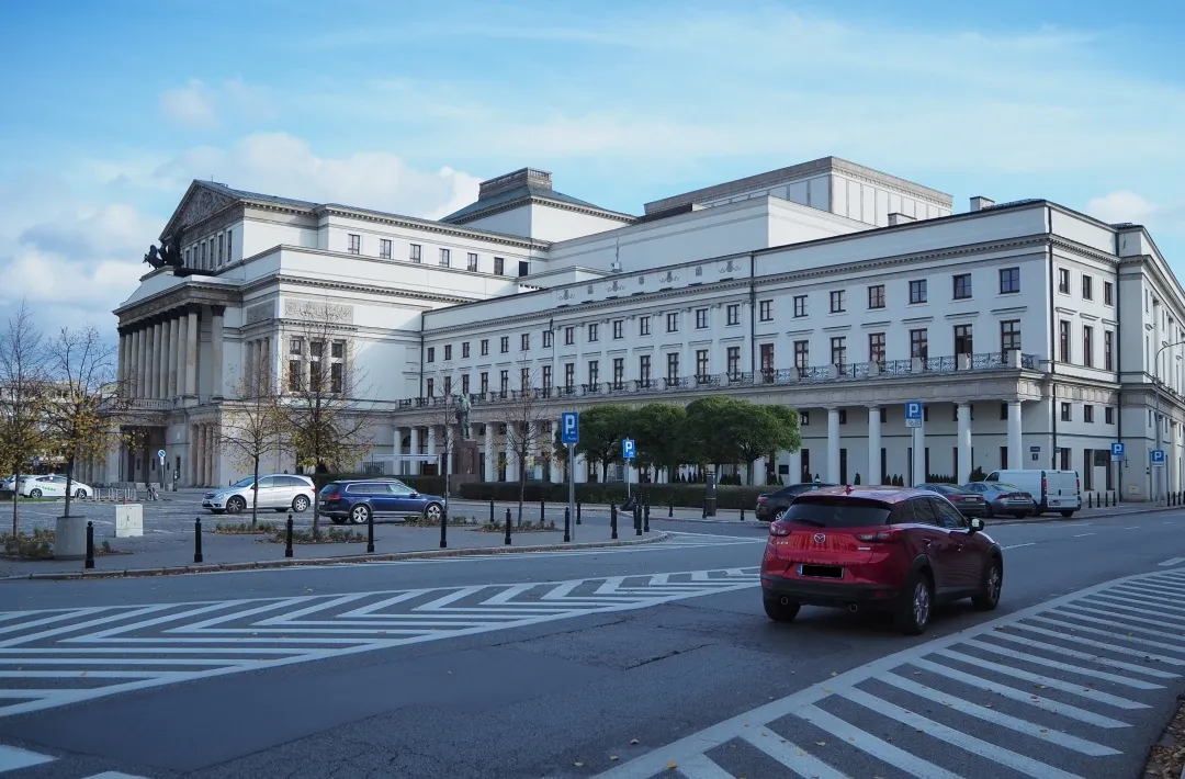 large white neoclassical building with columns and multiple windows, street with cars and crosswalk in foreground