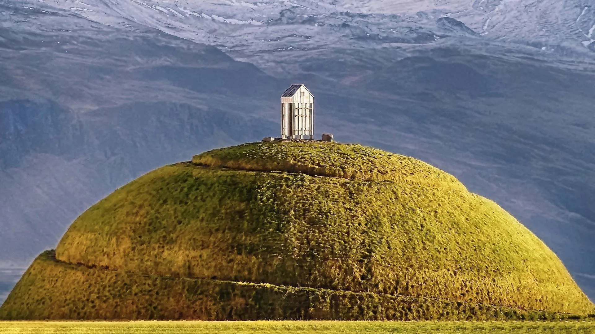Gran montículo en forma de cúpula cubierto de hierba con una pequeña estructura de madera blanca en la cima sobre un fondo montañoso.