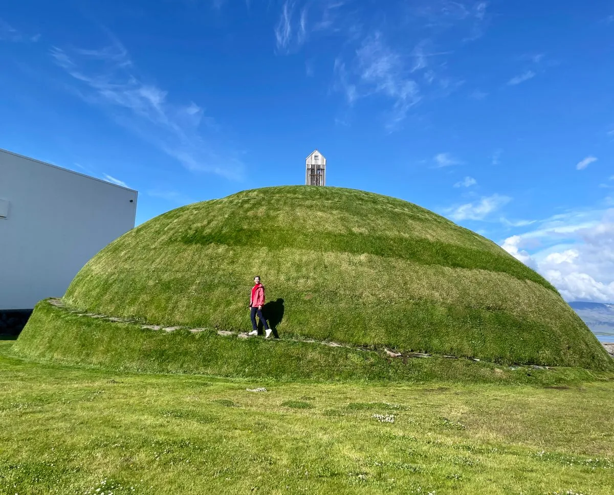 Montículo en forma de cúpula cubierto de hierba con un sendero en espiral y una persona de pie sobre él, coronado por una pequeña estructura blanca bajo un cielo azul