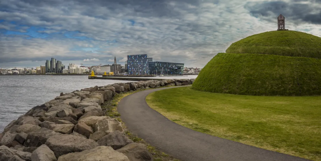 Un gran montículo cubierto de hierba con un sendero pavimentado que serpentea a lo largo de la costa rocosa del puerto, con el paisaje urbano de Reykjavík y la sala de conciertos Harpa visibles al otro lado del agua.