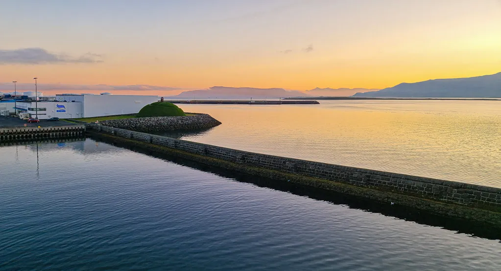 Colina cubierta de hierba al final de un muelle de piedra en el puerto de Reykjavík al atardecer, con aguas tranquilas que reflejan el cielo anaranjado y las montañas a lo lejos