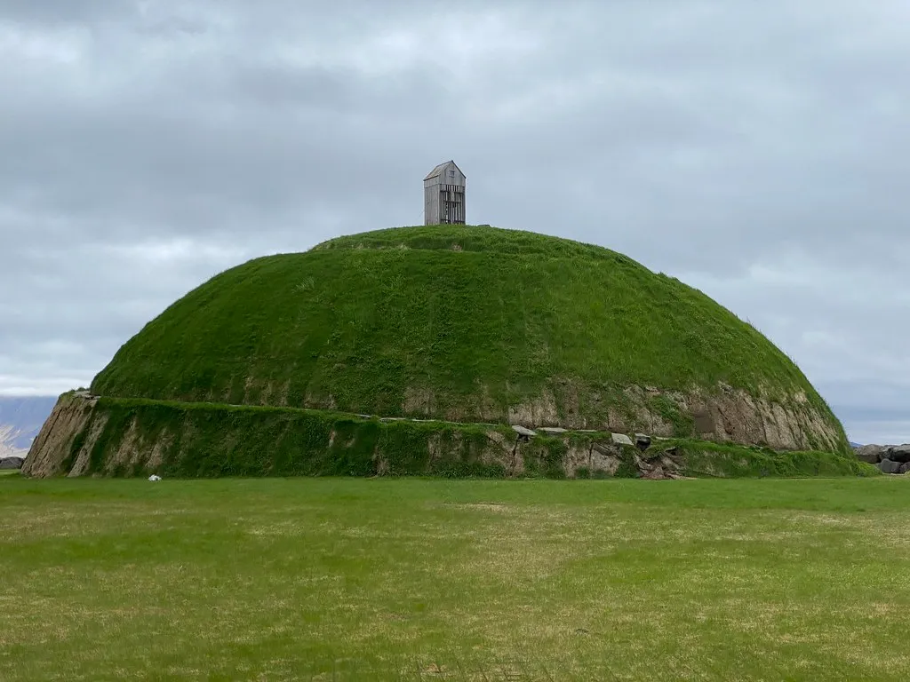 Gran montículo en forma de cúpula cubierto de hierba con una pequeña estructura de madera para secar pescado en la cima, rodeado de césped verde bajo un cielo nublado.