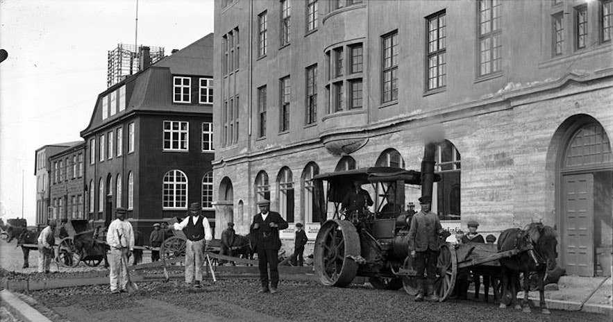 Black and white historical photograph of a street scene with a steamroller, horses, and workers in front of large brick buildings in the Hafnartorg district.