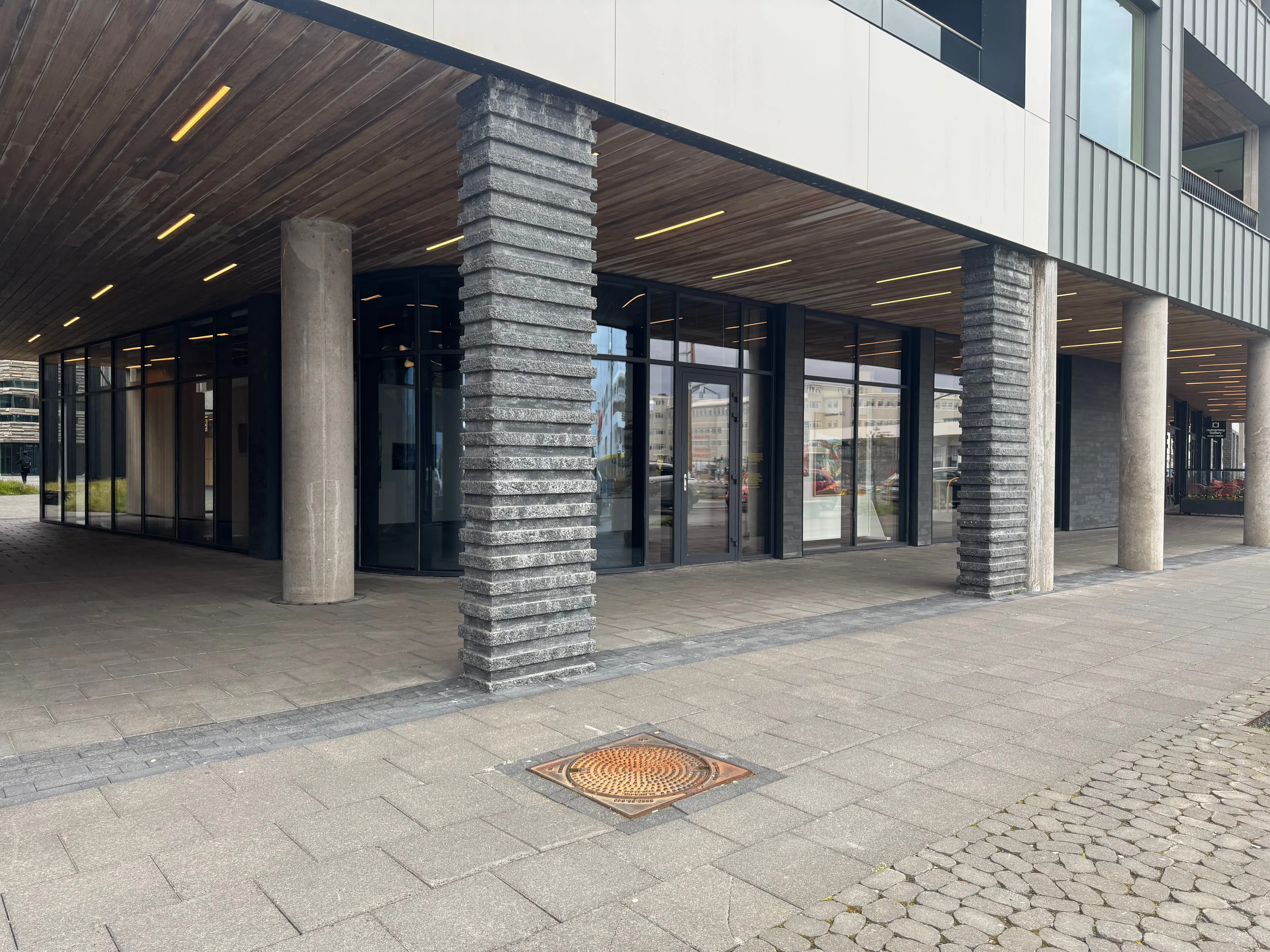 Modern building exterior with glass storefront windows, concrete and dark stone-clad pillars, and a wooden ceiling overhang above a paved walkway