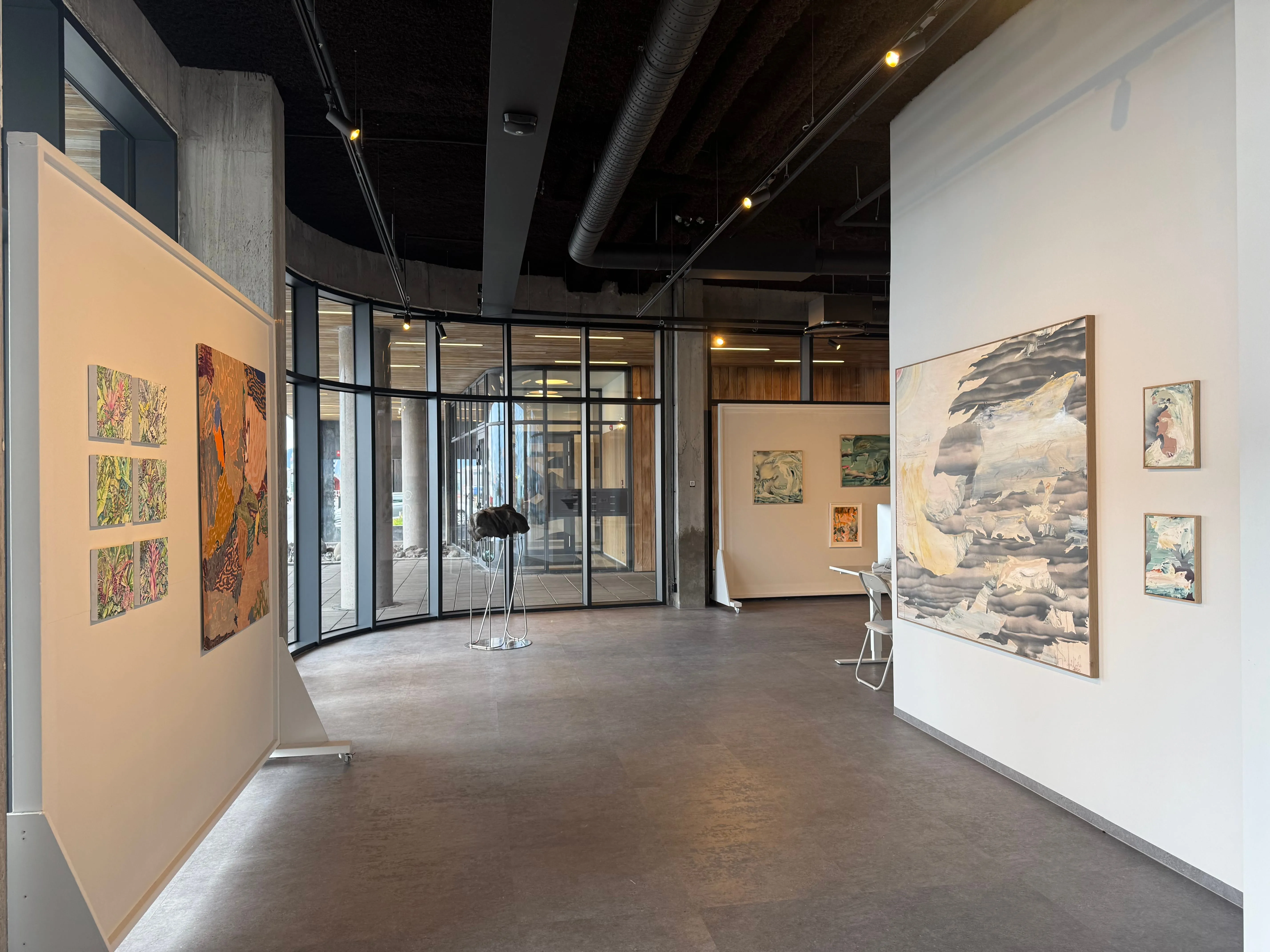 Interior view of Þula gallery showing paintings on white walls, curved glass entrance, exposed ceiling with track lighting, and polished concrete floor