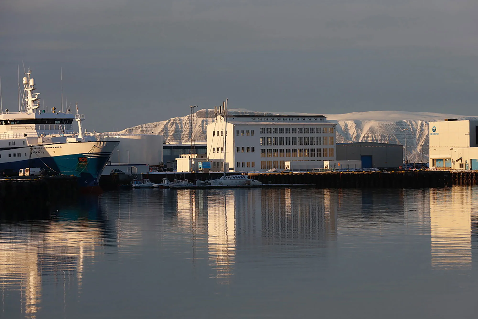 Widok na port z dużym statkiem zacumowanym przy białych budynkach przemysłowych, z ośnieżoną górą w tle i spokojną wodą, w której odbijają się budowle.