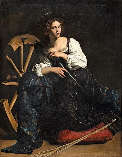 Een vrouw in een witte blouse en donkere rok zit naast een houten wiel, met een staf in haar hand, tegen een donkere achtergrond en met dramatische belichting.