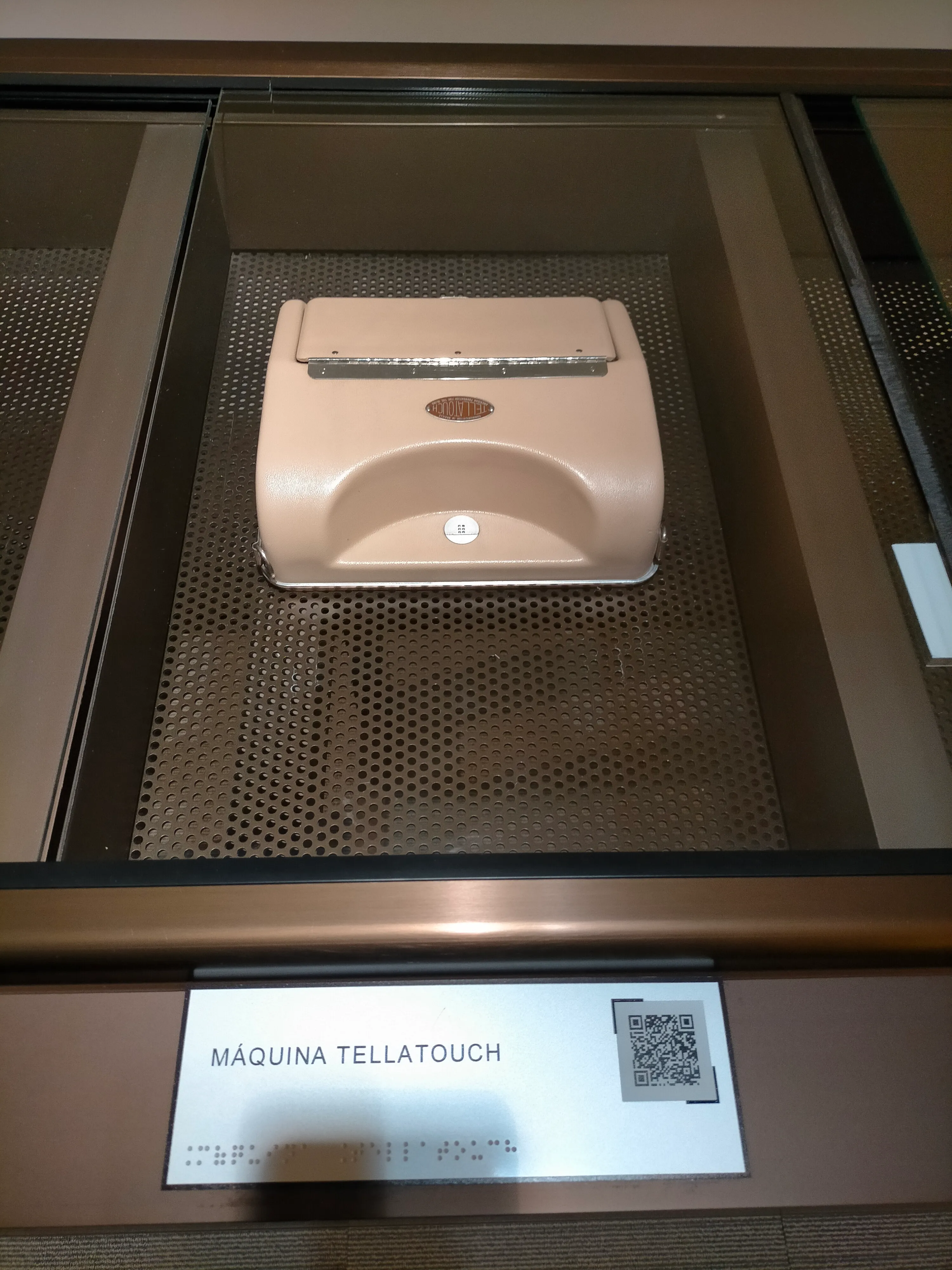 Una máquina táctil beige etiquetada como 'MÁQUINA TELLATOUCH' dentro de una vitrina de cristal con una base de metal perforado.